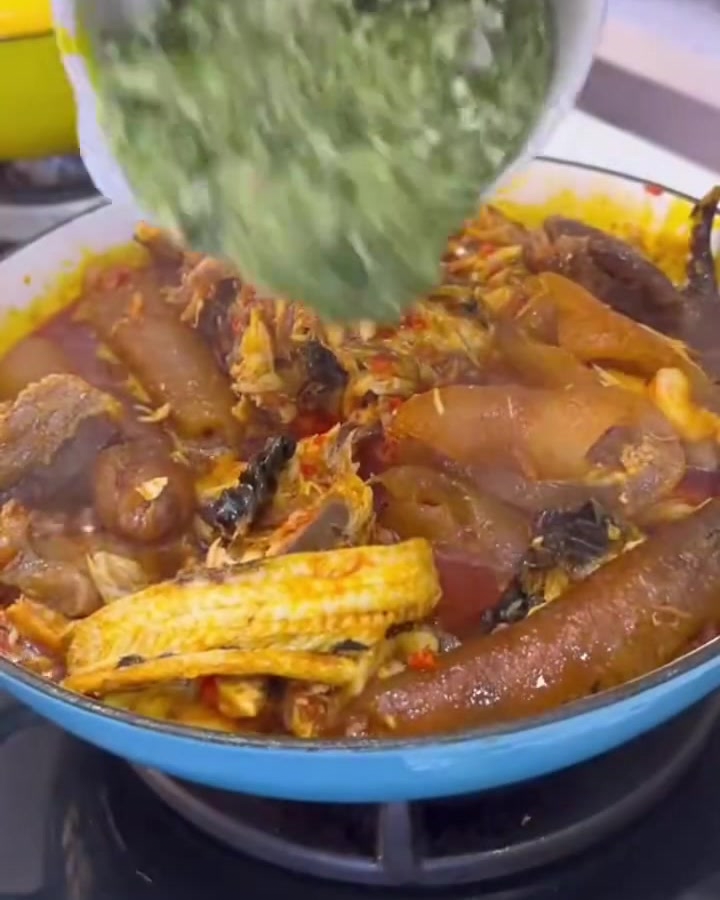 Finely chopped green okra being poured from a white bowl into a rich, bubbling pot of meats and sauce.
