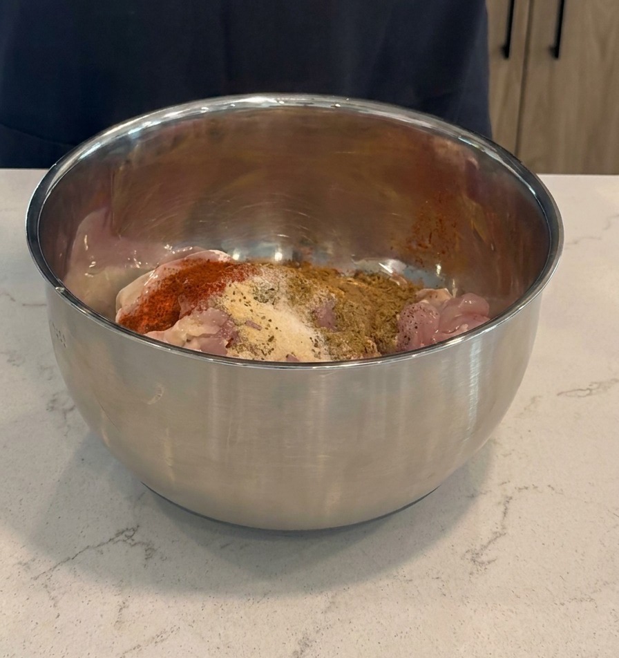Adding dark red marinade and dry spices to raw chicken thighs inside a stainless steel mixing bowl.