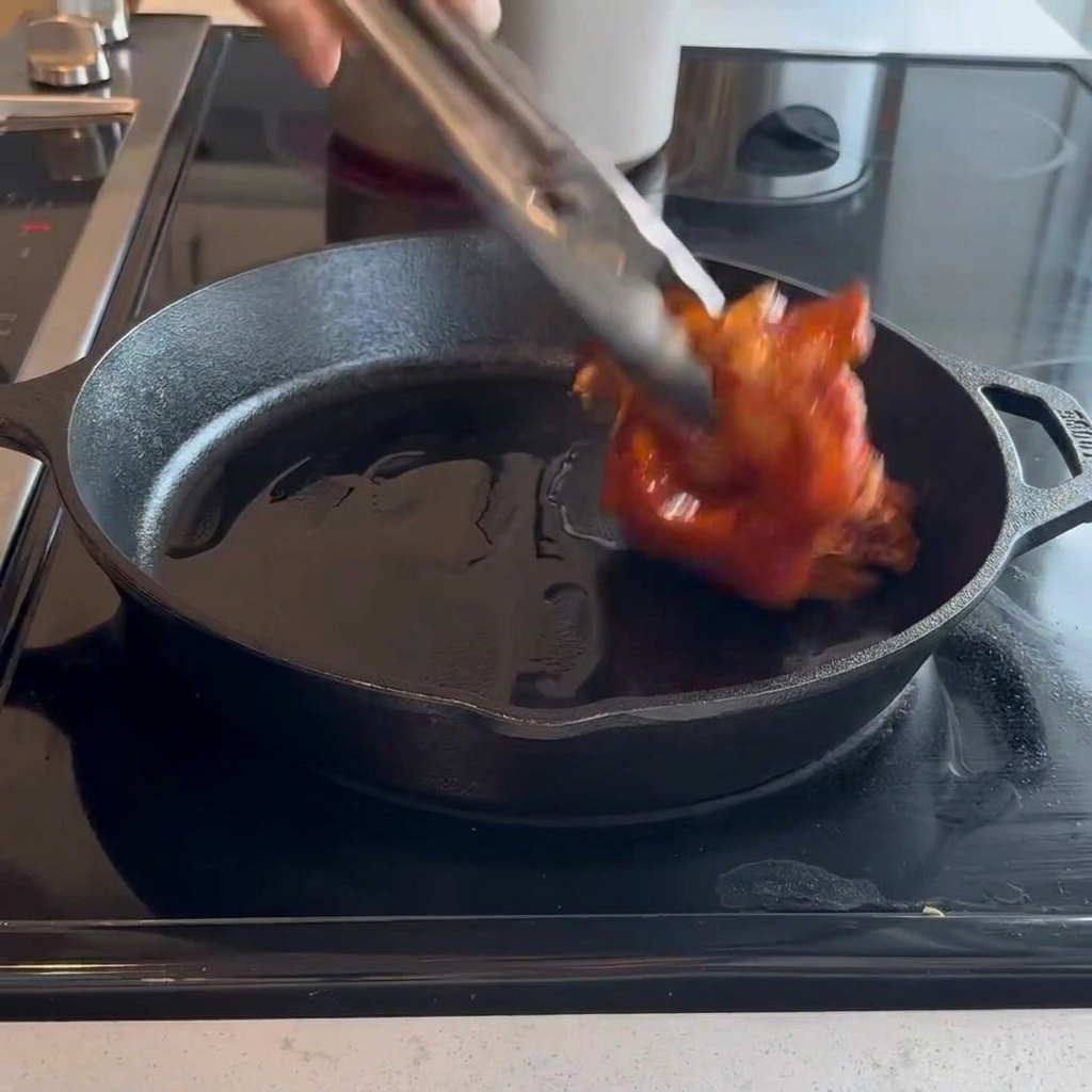 Raw marinated chicken thighs being placed skin-side down into a hot cast-iron skillet using metal tongs.