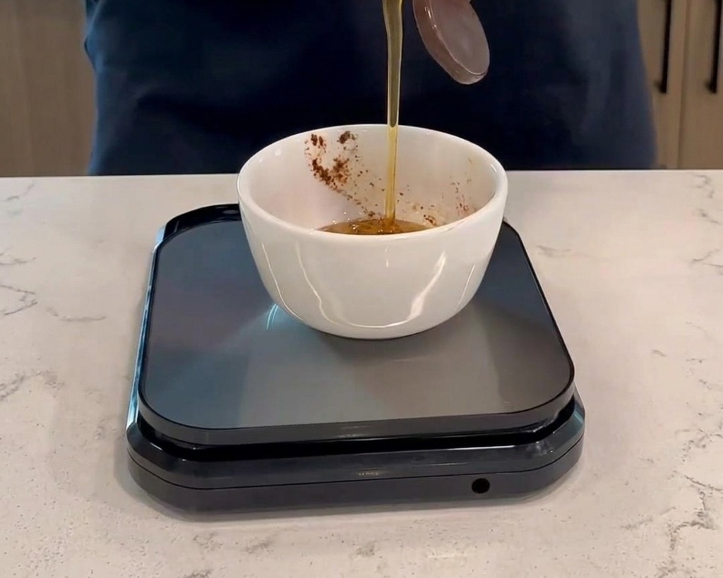 Pouring honey from a plastic bottle into a small white bowl with dark red sauce, placed on a black digital kitchen scale.