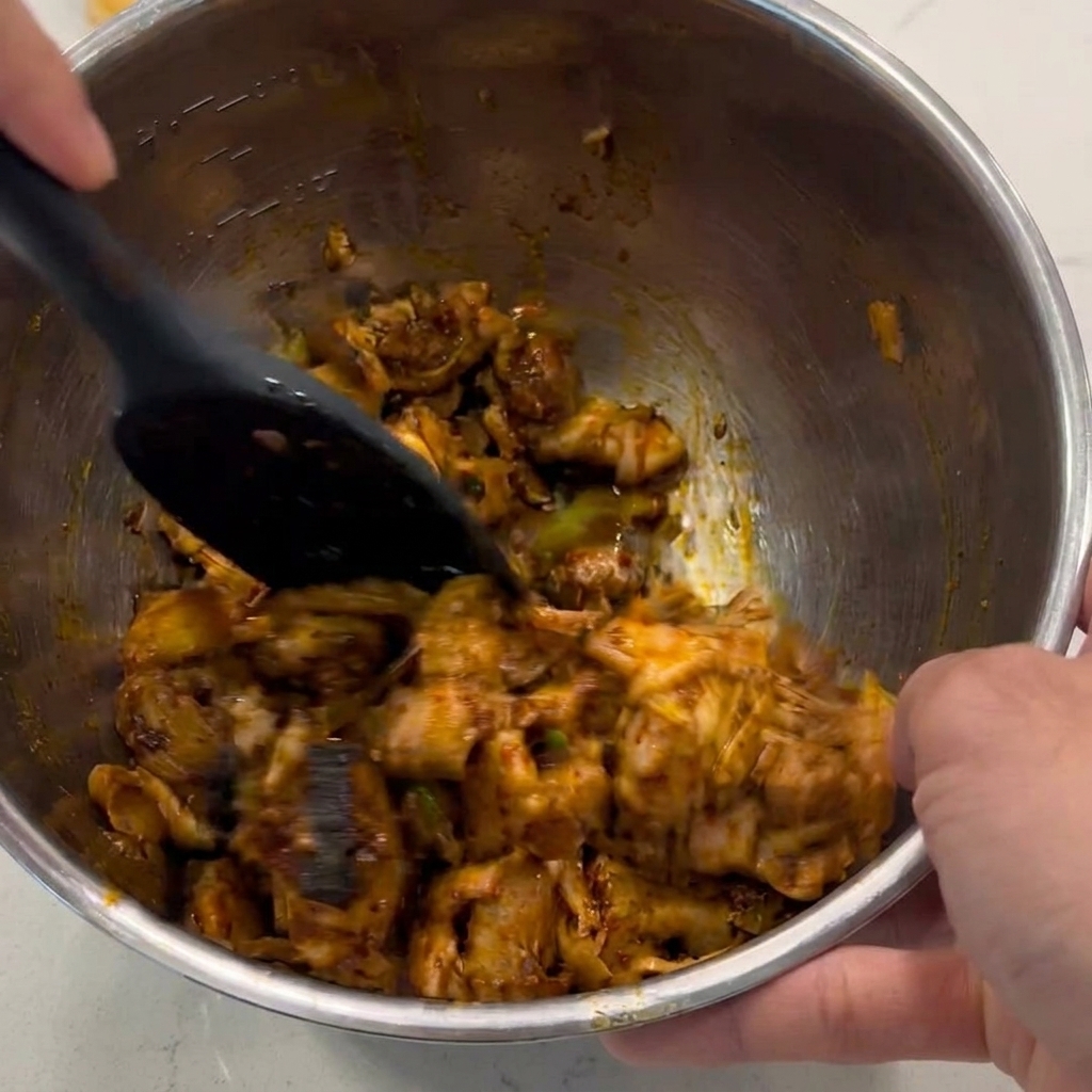 A black spatula mixing and stirring diced chicken chunks to coat them in a glossy brown sauce inside a metal bowl.