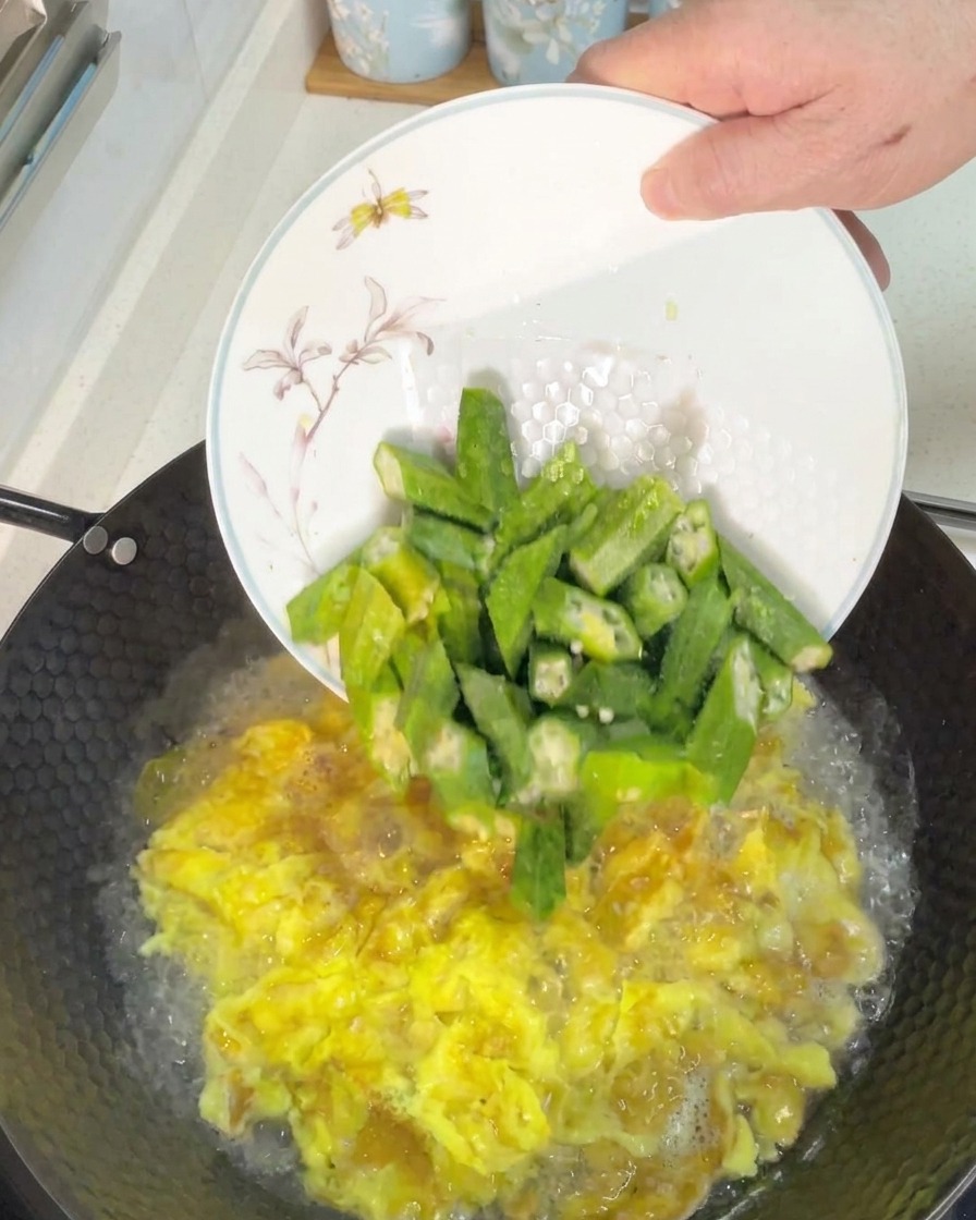 Adding freshly sliced green okra pieces into a wok of bubbling egg soup.