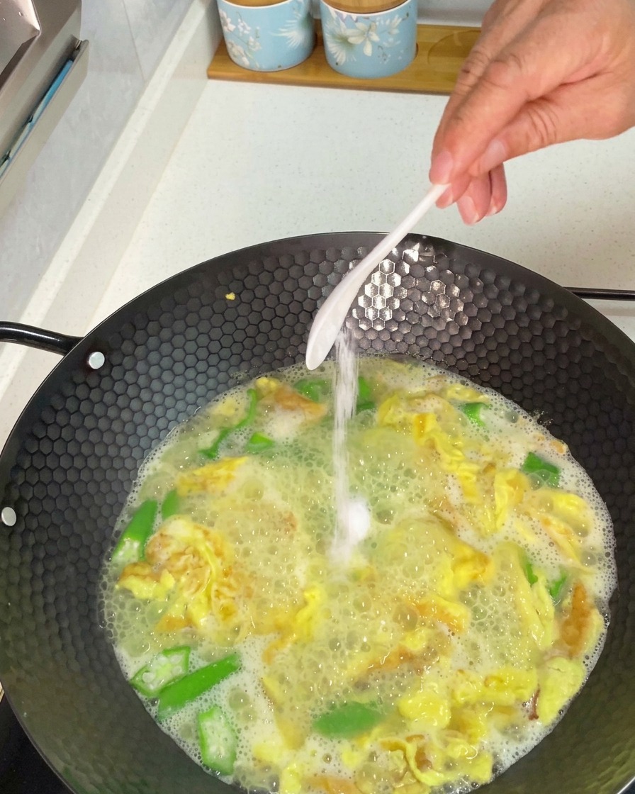 Hand adding a spoonful of white salt to a simmering wok of okra and egg soup.
