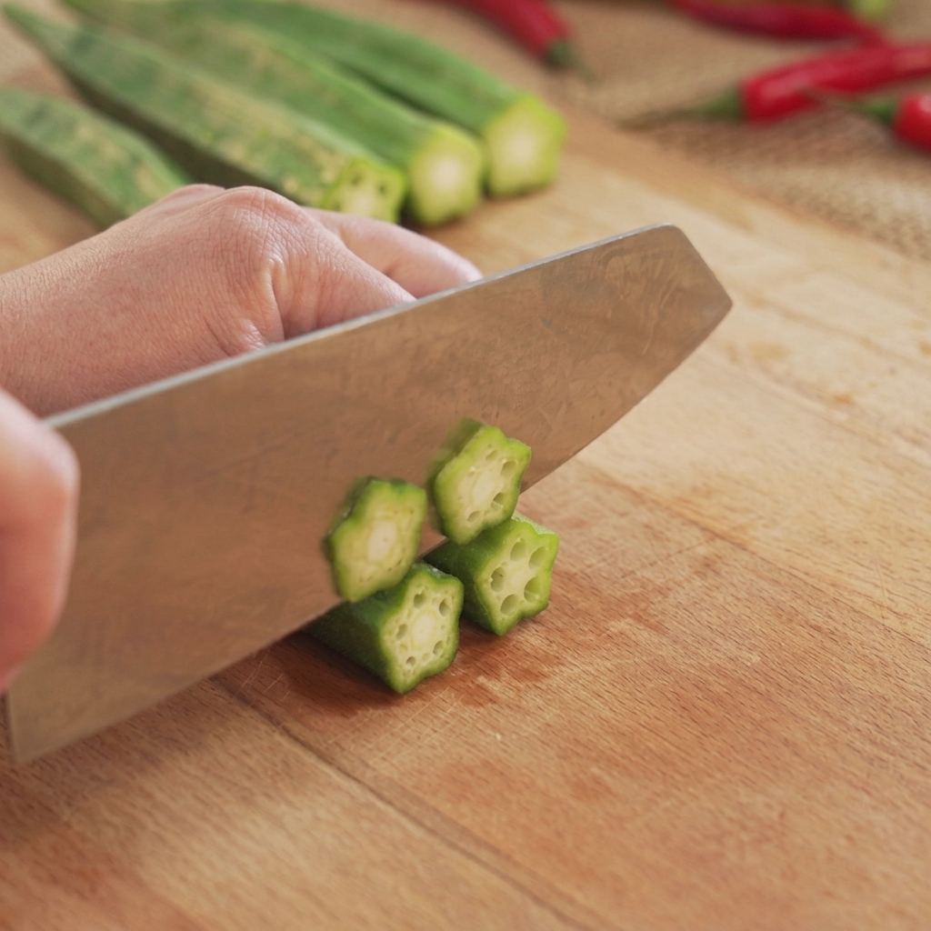 Green okra being cut into round star-shaped slices on a wooden chopping board with a kitchen knife.
