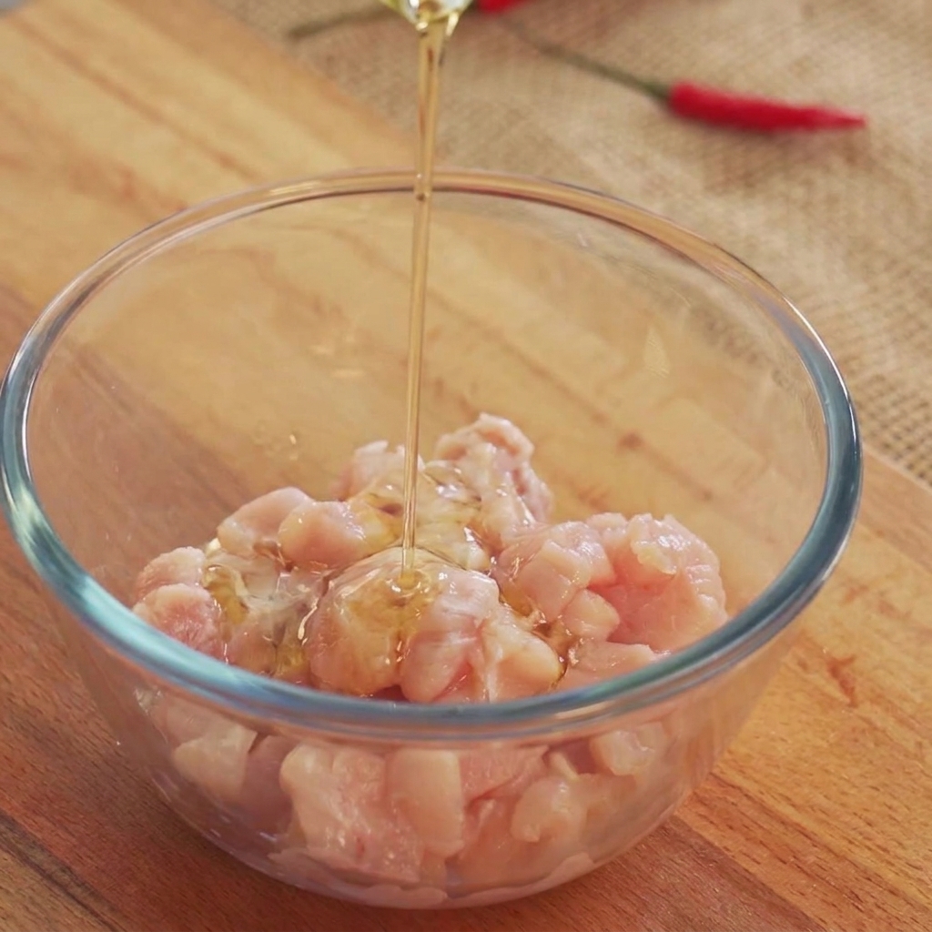 Cooking oil being poured from above onto raw chicken chunks inside a glass bowl.