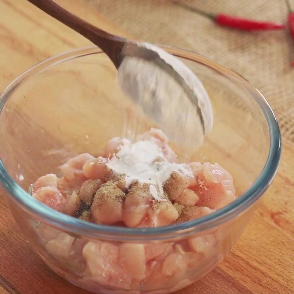 Cooking wine and white seasoning powders being sprinkled onto raw chicken cubes in a glass bowl.
