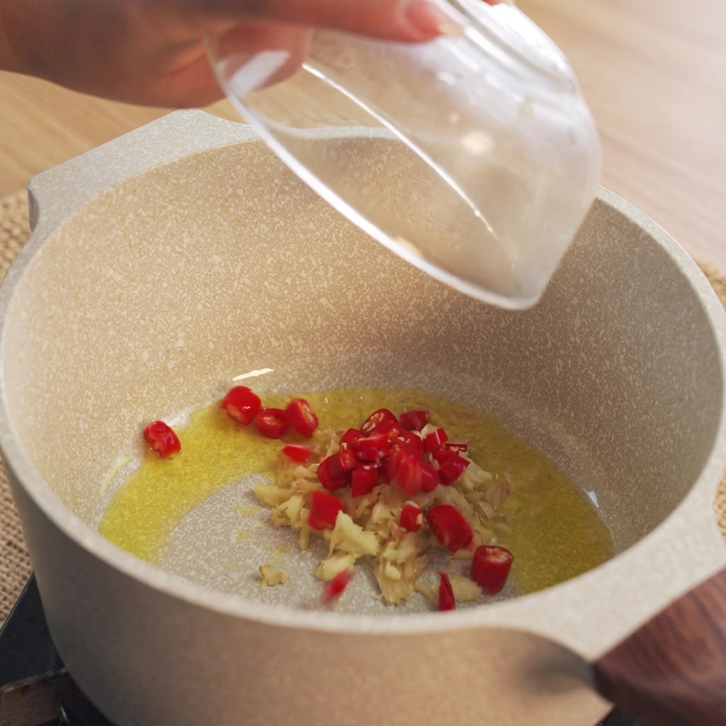 Hand dropping chopped red chili peppers into a pot with heated oil and minced ginger.