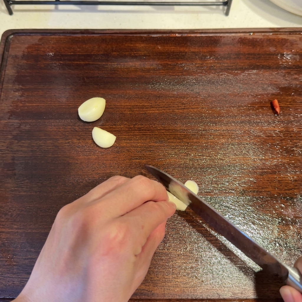Garlic cloves being sliced with a knife on a wooden cutting board next to a red chili piece.