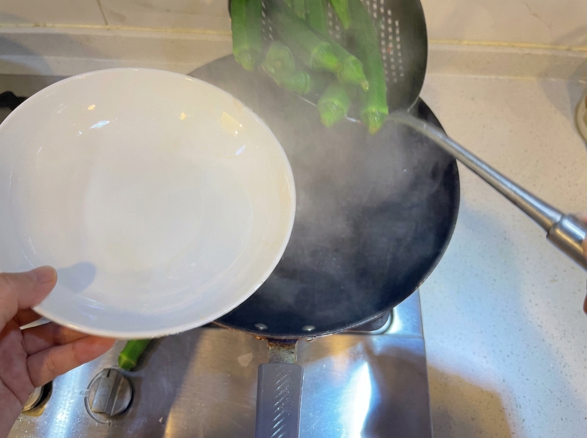 Whole green okra being blanched inside a pan of boiling water using a strainer ladle.