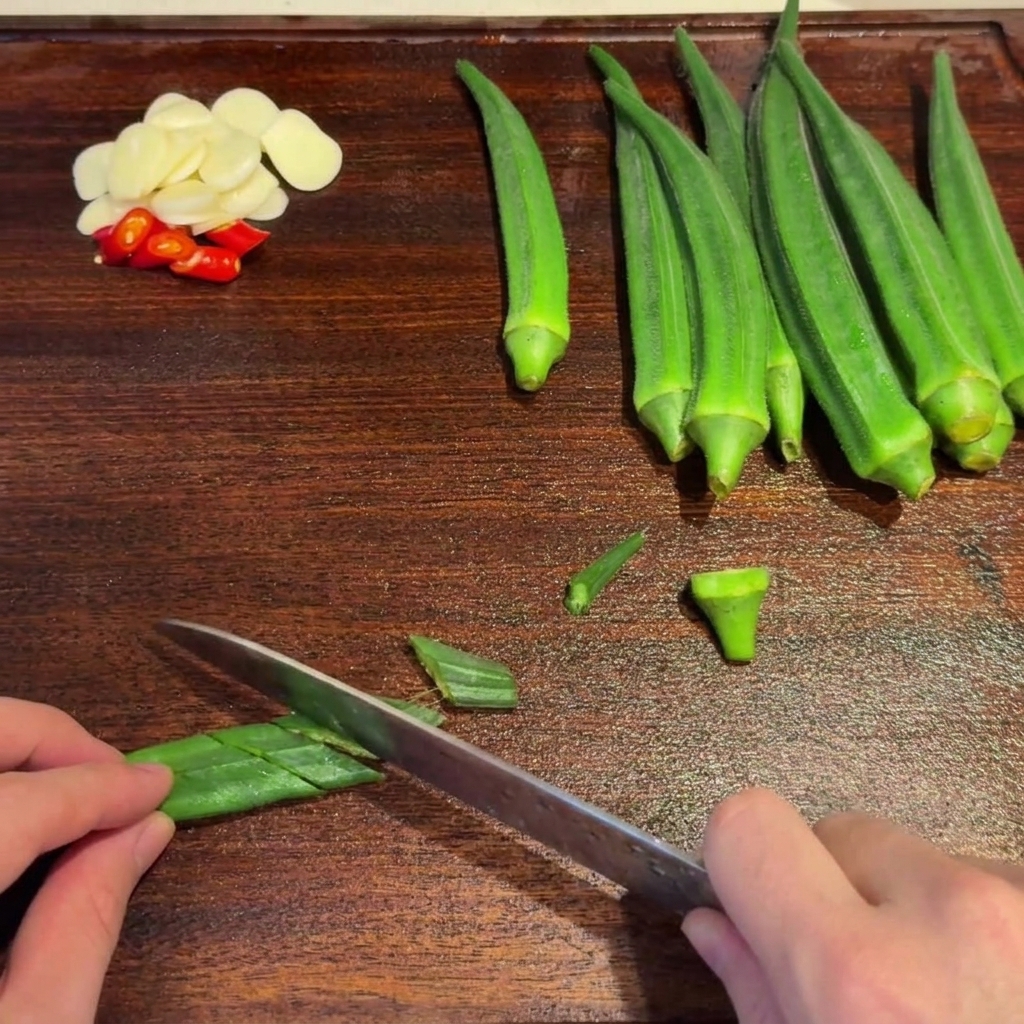 Blanched green okra being sliced diagonally with a knife on a wooden cutting board.