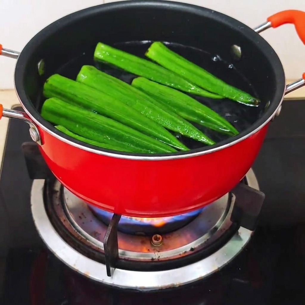 Raw whole okra pods being placed into a red pot of boiling water.