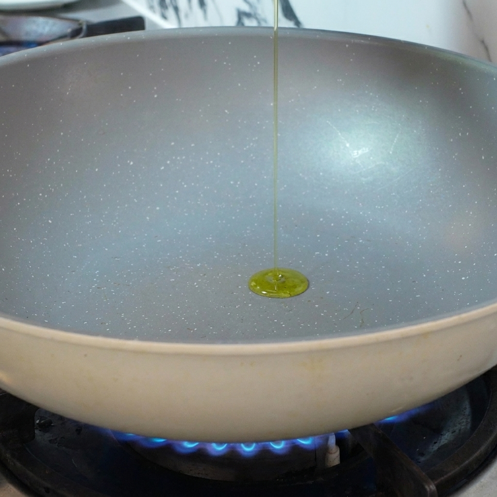 Olive oil being poured into a preheated ceramic frying pan over a blue gas flame.