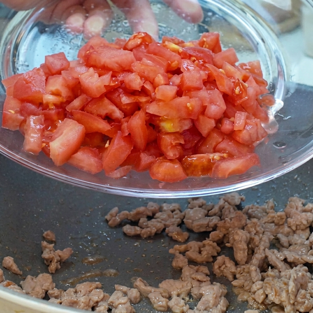 Diced tomatoes and red onions being poured from a glass bowl into the pan with the meat.