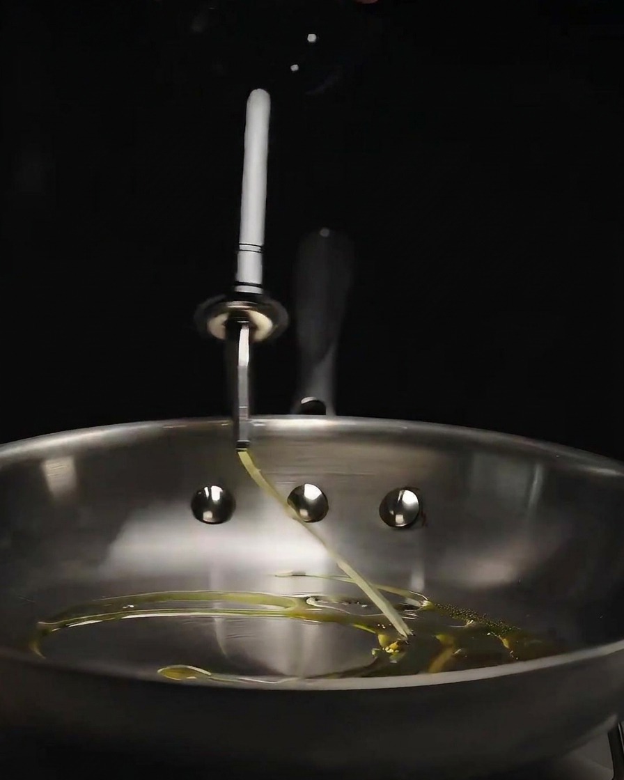 Olive oil being poured from a glass bottle into a shiny stainless steel frying pan.