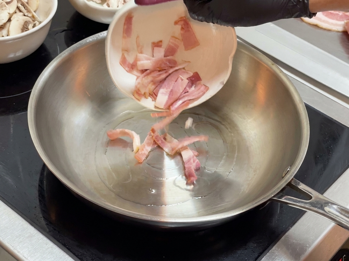 Bacon pieces being poured from a small white bowl into a stainless steel frying pan.