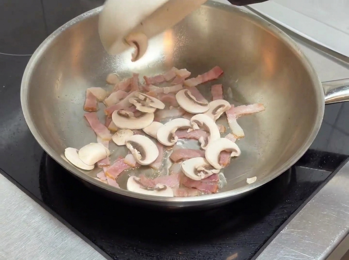 Sliced mushrooms being added to the pan containing cooked bacon pieces.