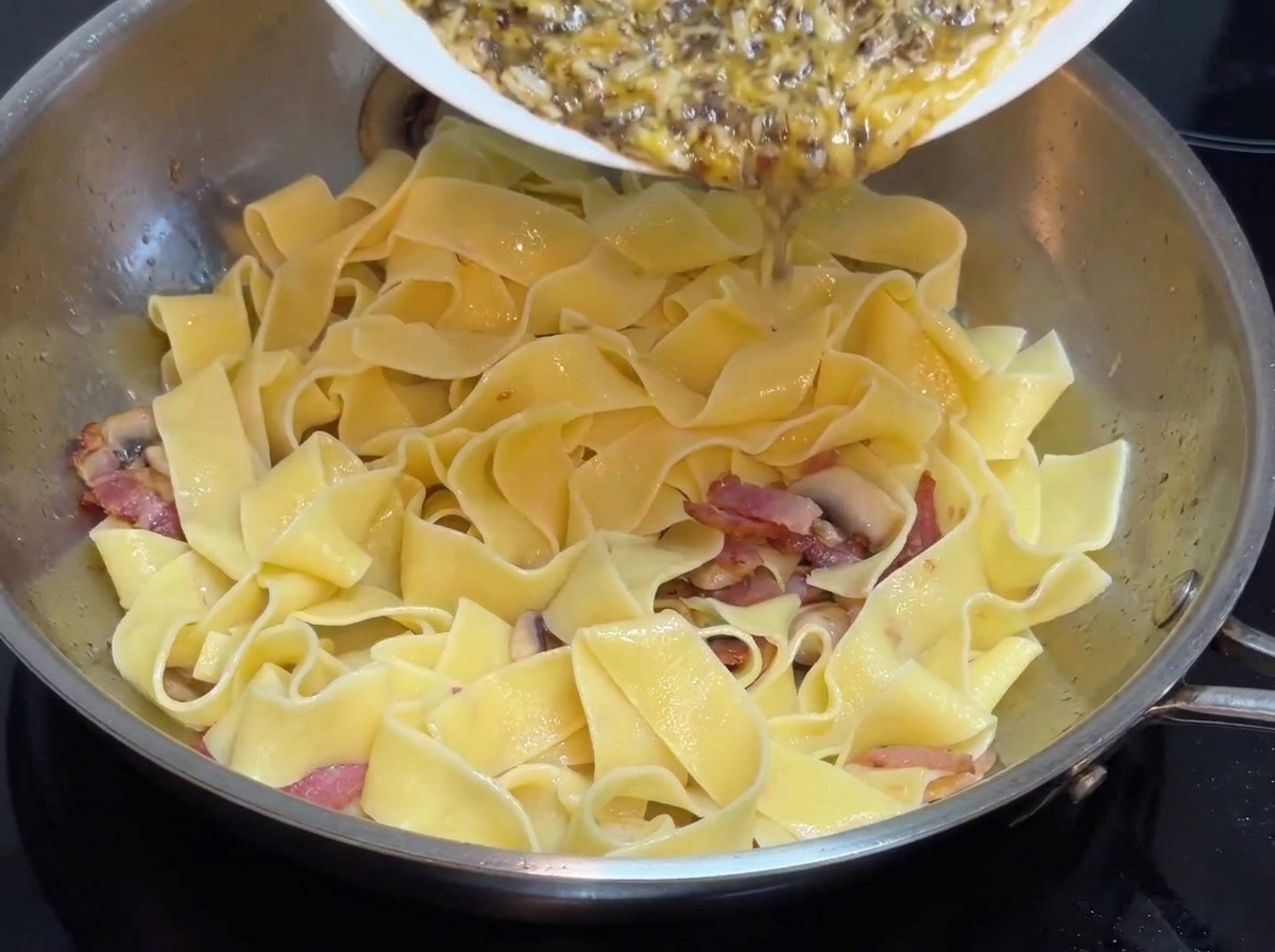 Pre-made black truffle custard sauce being poured from a white plate into a stainless steel pan containing wide pasta.