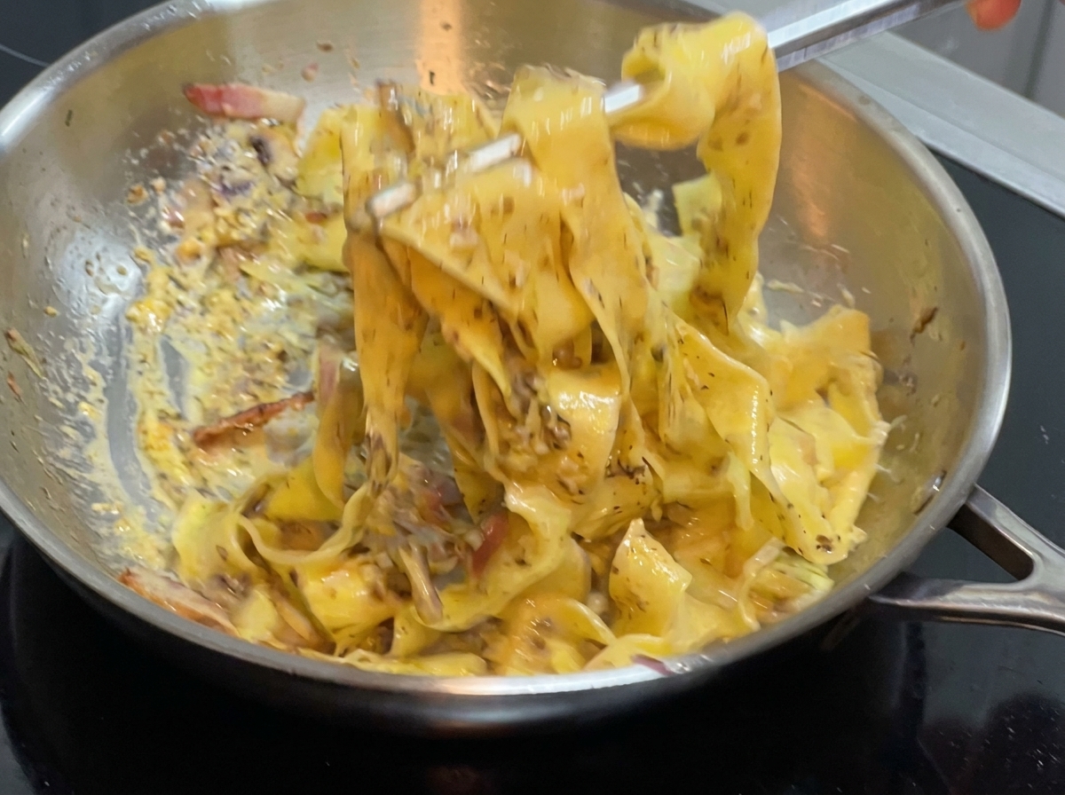A pair of tongs mixing wide pasta and sauce inside a stainless steel pan on the stove.
