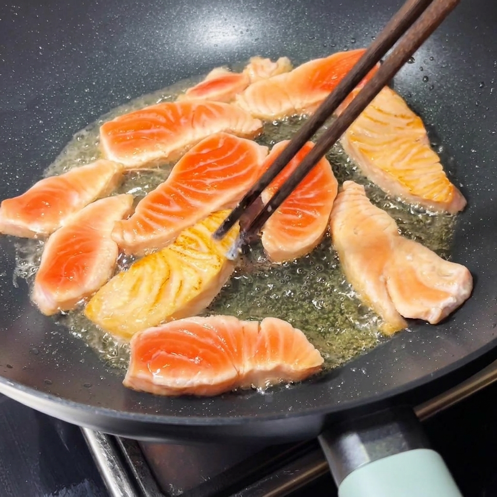 A pair of wooden chopsticks flipping a golden-brown seared salmon piece inside a hot frying pan.