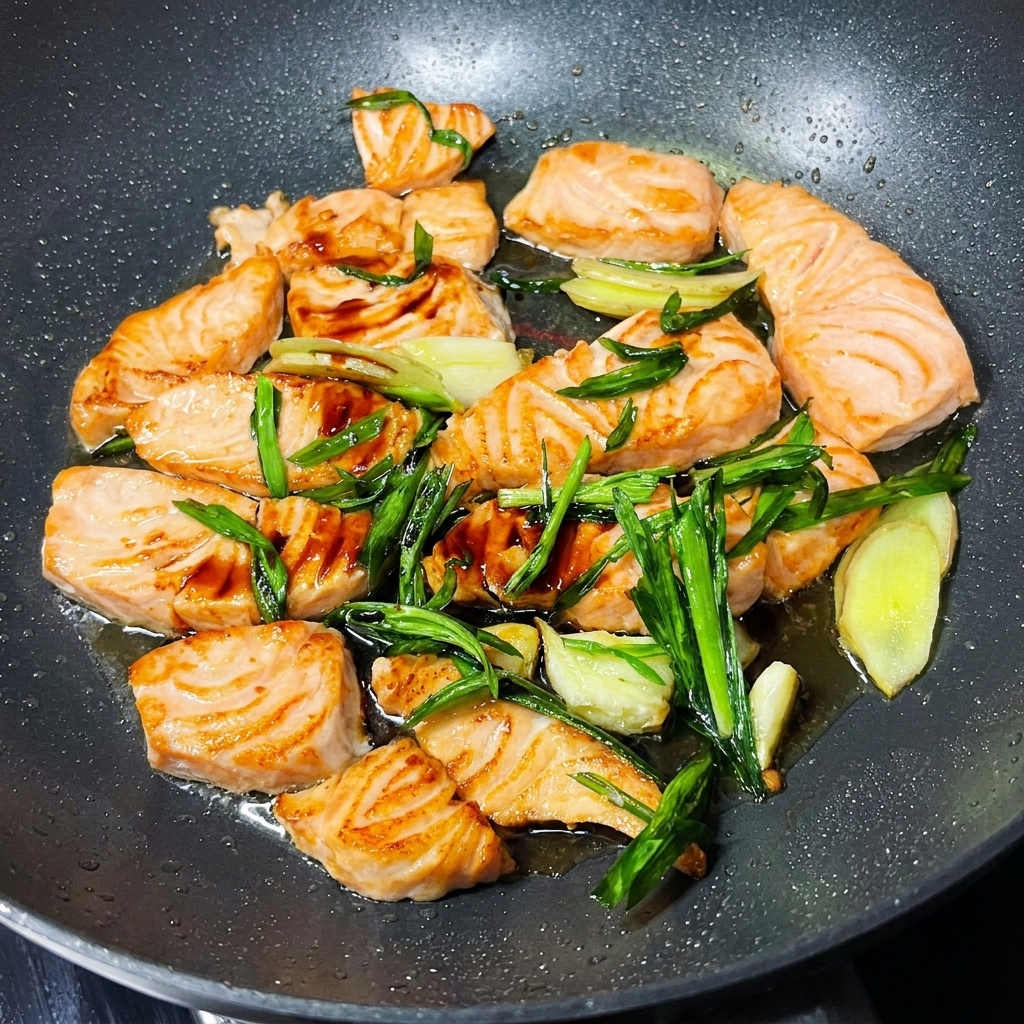 Salmon pieces in a pan coated with soy sauce and garnished with fresh green scallions and sesame seeds.