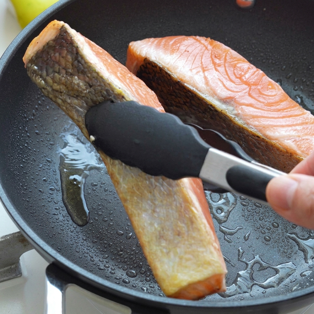Salmon fillets being flipped with black tongs inside a hot skillet with oil, showing golden-brown seared skin.