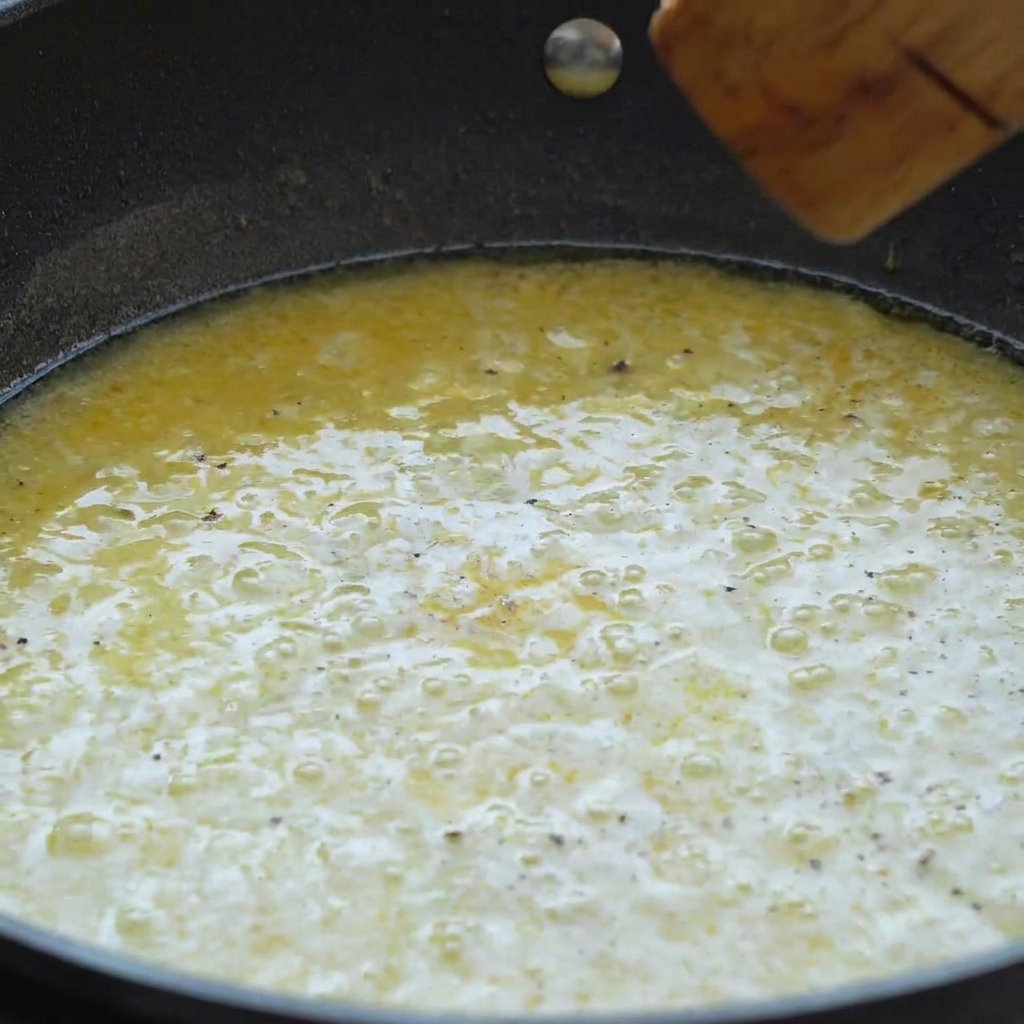 A pan filled with a simmering creamy yellow lemon butter sauce being mixed with a wooden spatula.