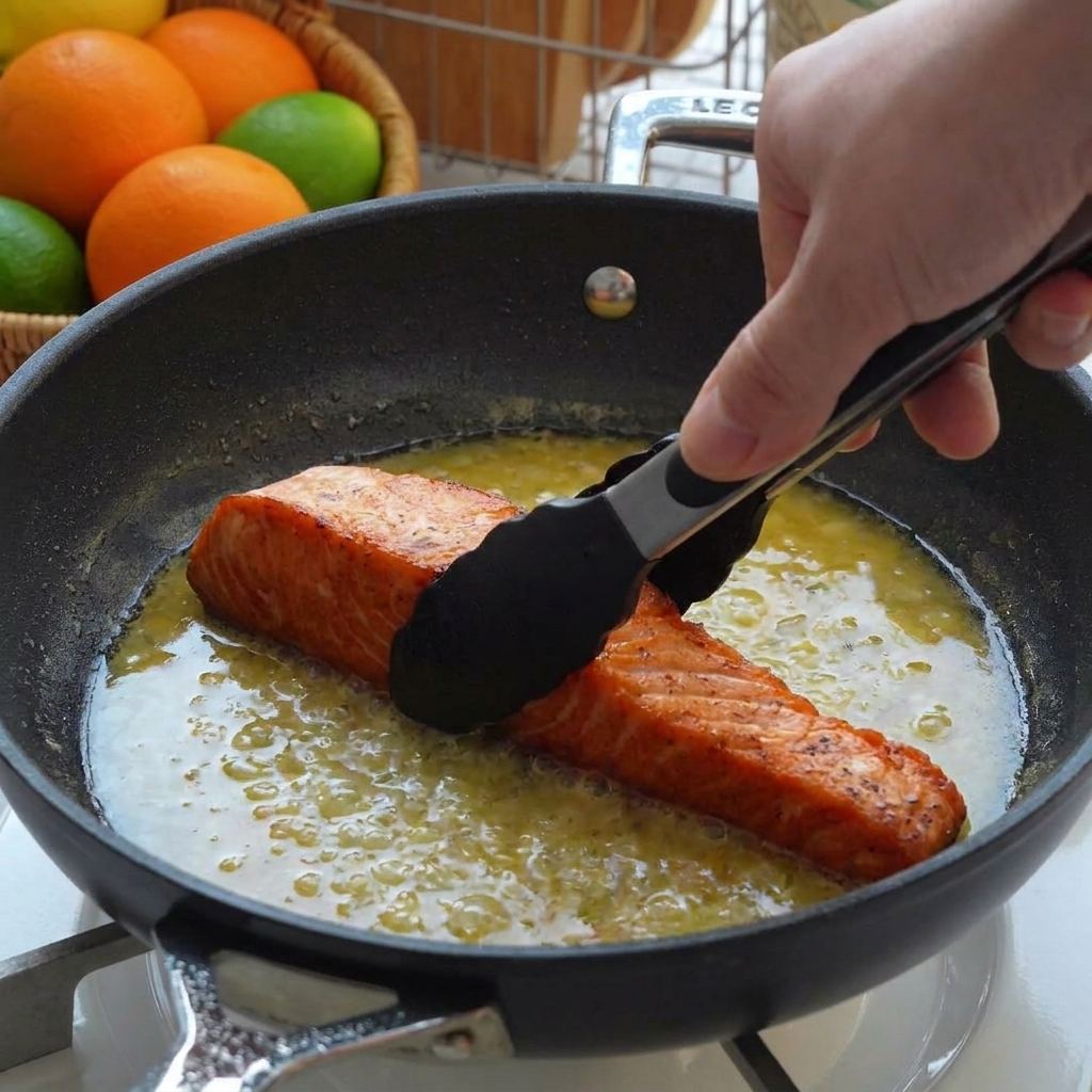 Seared salmon fillets being placed into a pan filled with lemon butter sauce using black tongs.
