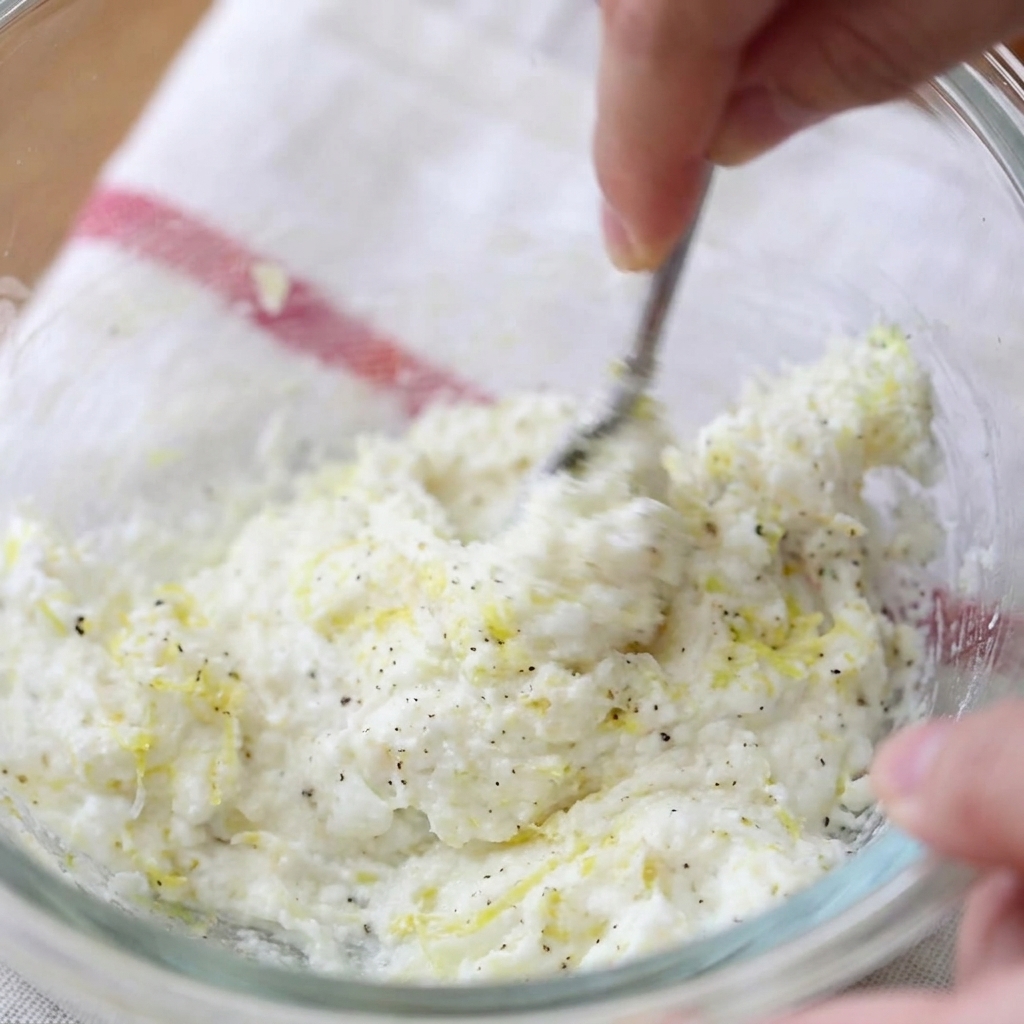 A hand using a fork to mix cream cheese, lemon zest, and black pepper in a glass bowl.