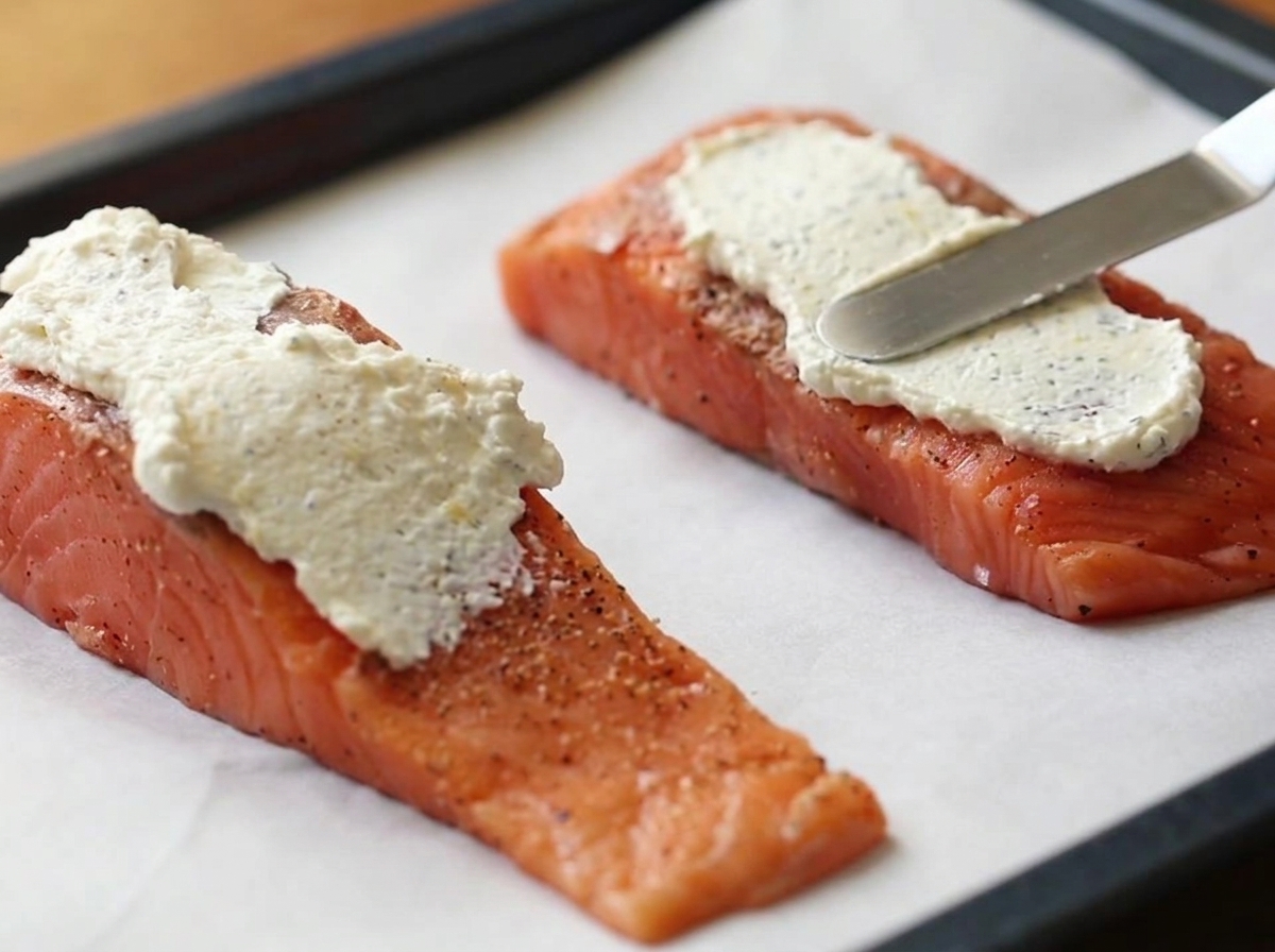 A metal spatula spreading a thick layer of white cream cheese mixture onto two raw salmon fillets on a parchment-lined baking tray.