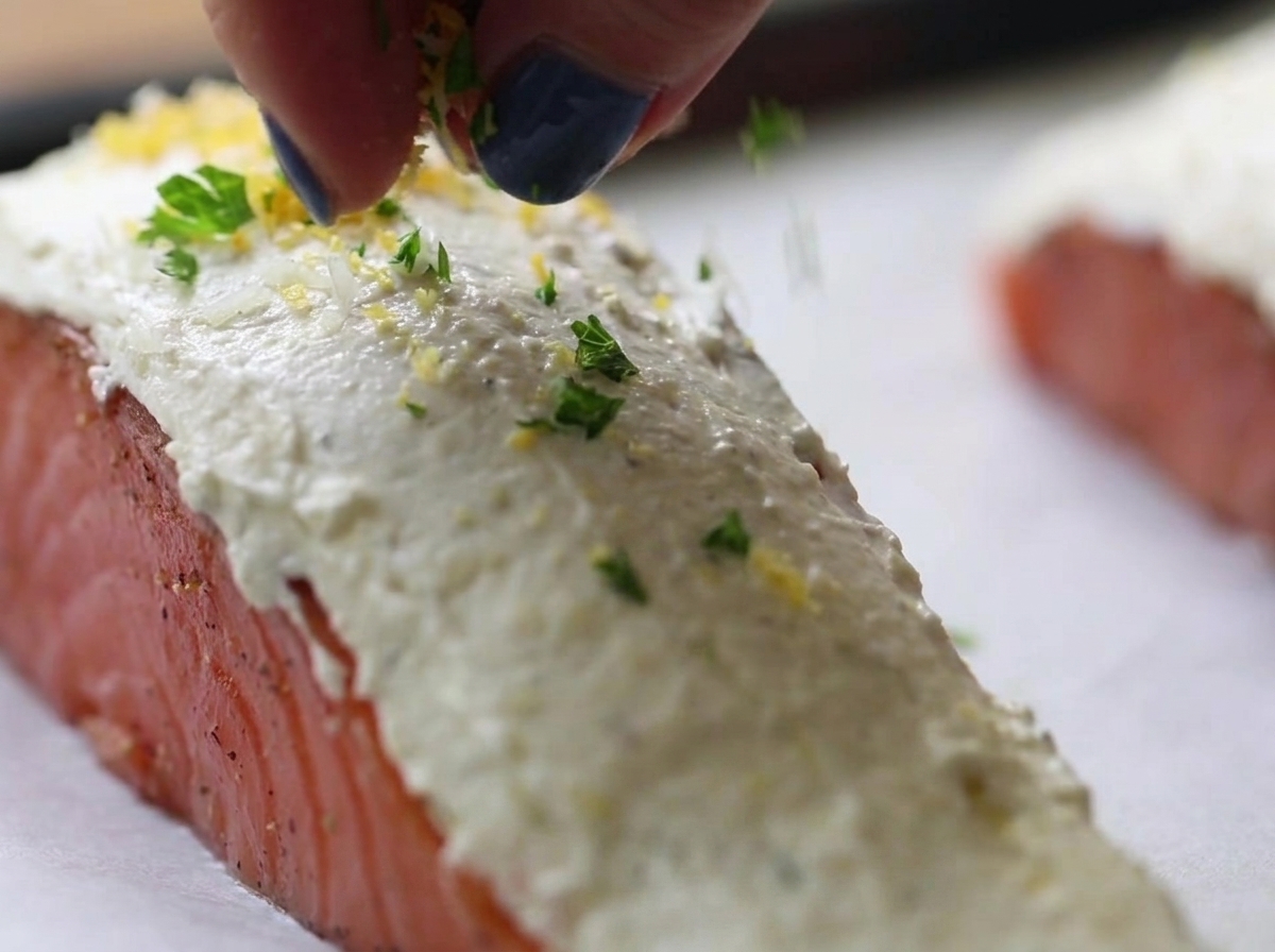 Fingers sprinkling a green and yellow herb breadcrumb mixture over raw salmon fillets coated in white cream cheese.