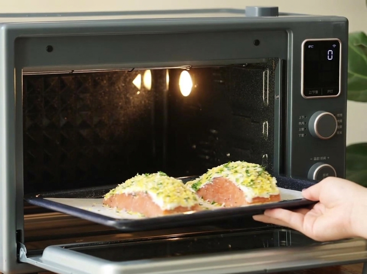 A hand sliding a dark baking tray with two breaded salmon fillets into a small, brightly lit countertop oven.
