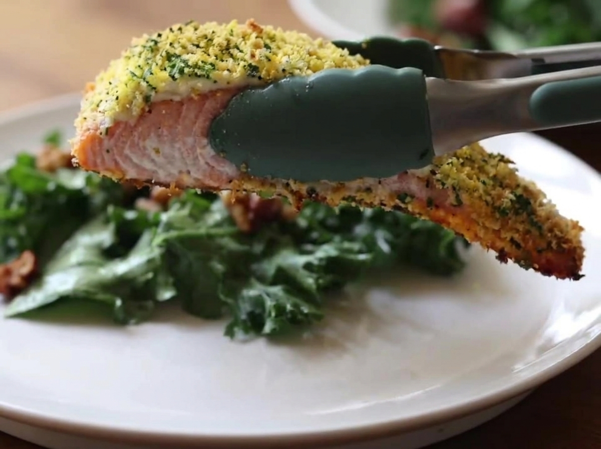 Green silicone kitchen tongs holding a piece of baked salmon with a golden, crispy crust over a white plate containing a green kale salad.