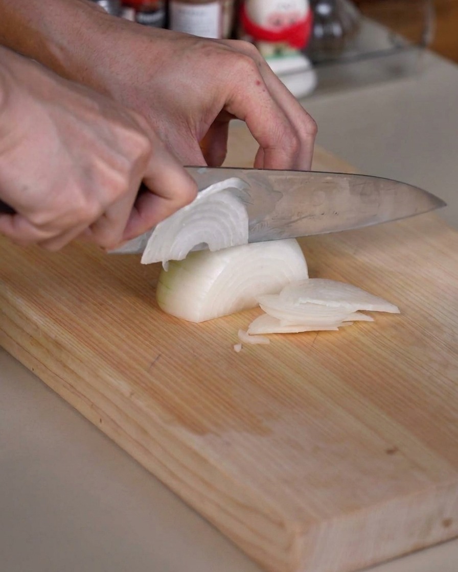 An onion being carefully sliced into thin pieces on a wooden cutting board using a sharp kitchen knife.