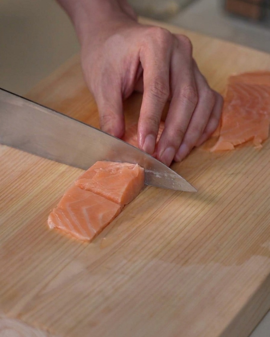 Fresh salmon fillet being portioned into small bite-sized cubes on a wooden cutting board.