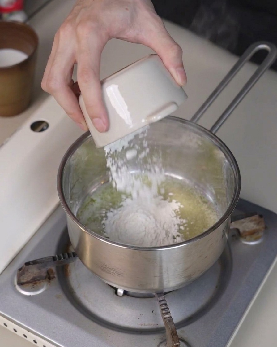 Flour being poured from a small bowl into melted butter in a saucepan, being stirred with a spatula.