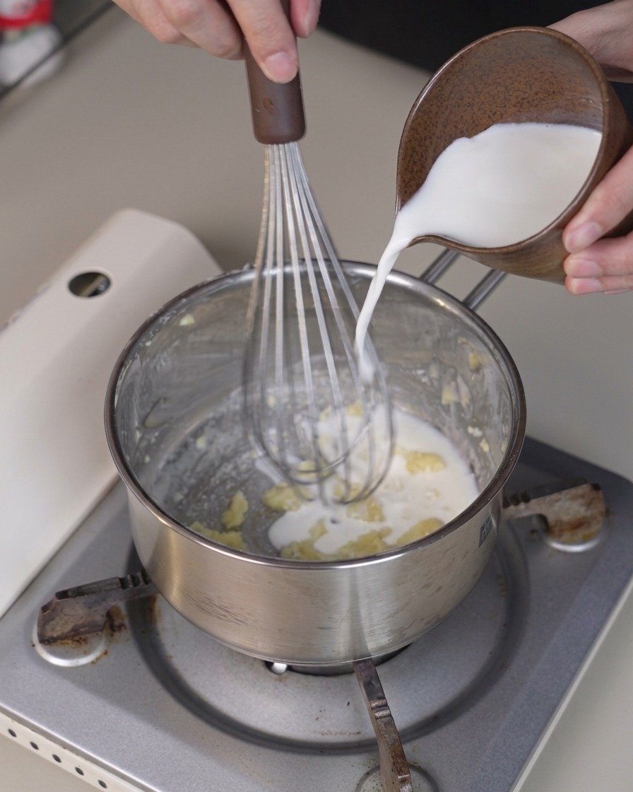 Cold milk being poured from a small pitcher into a saucepan while a whisk stirs the mixture.
