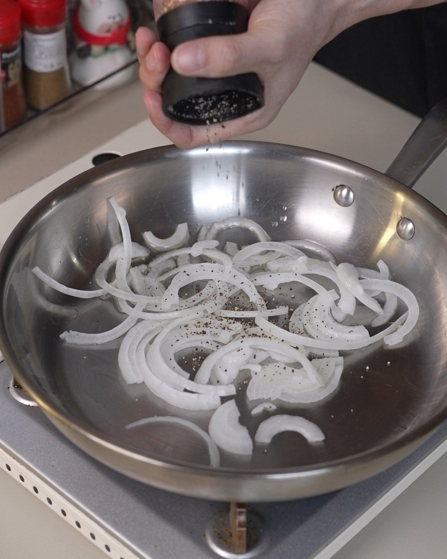 Sliced onions being seasoned with pepper and tossed in a stainless steel frying pan.