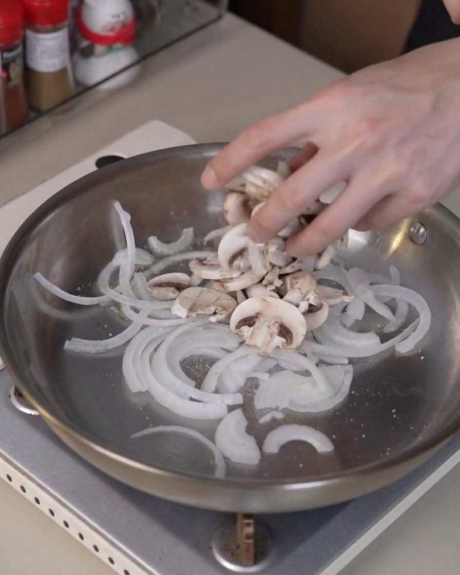 Sliced button mushrooms being added into the pan with onions to cook and release moisture.