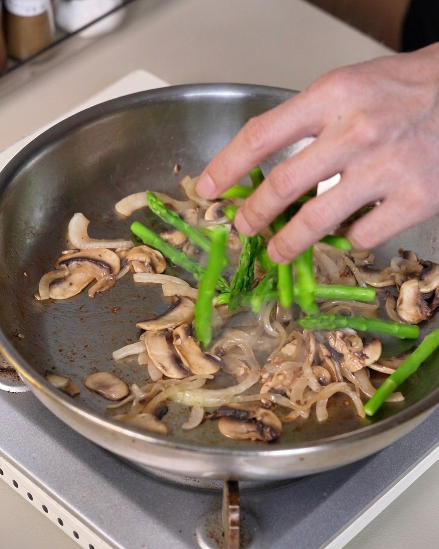 Green asparagus pieces being mixed into the pan containing sautéed onions and mushrooms.