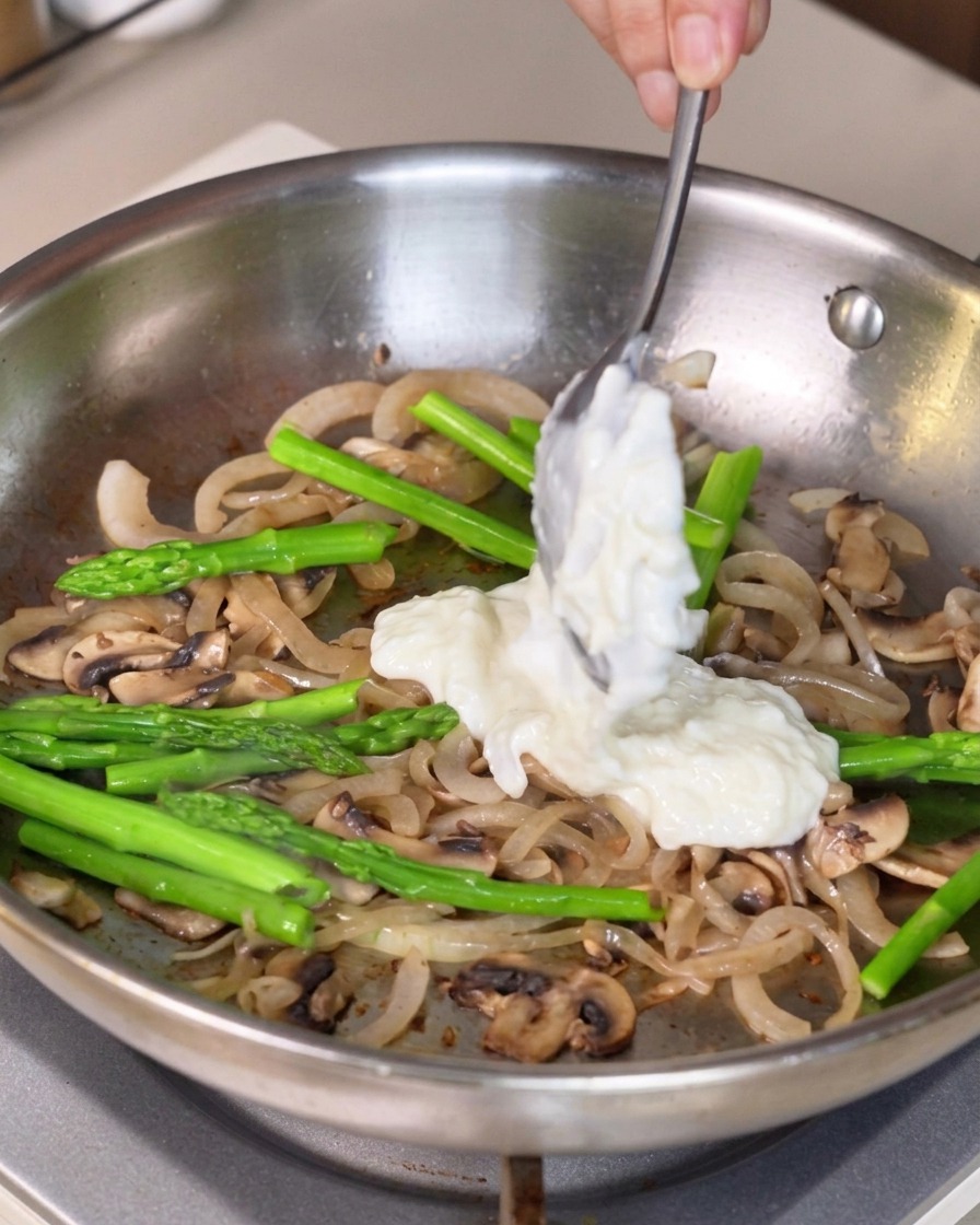 Thick white sauce being scooped with a spoon and added to the vegetable mixture in the pan.