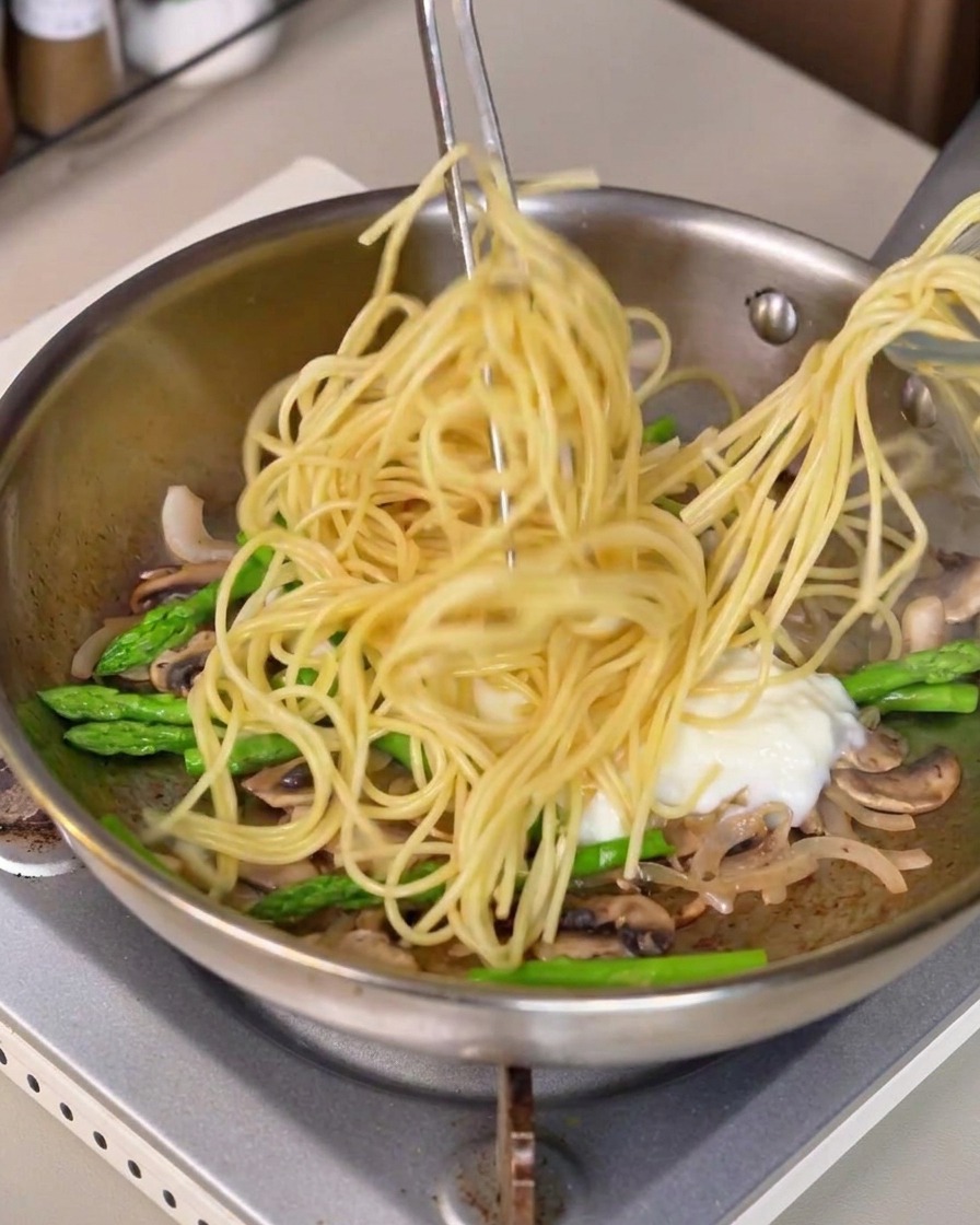 Long spaghetti pasta being lifted with tongs and mixed into the pan with white sauce and vegetables.