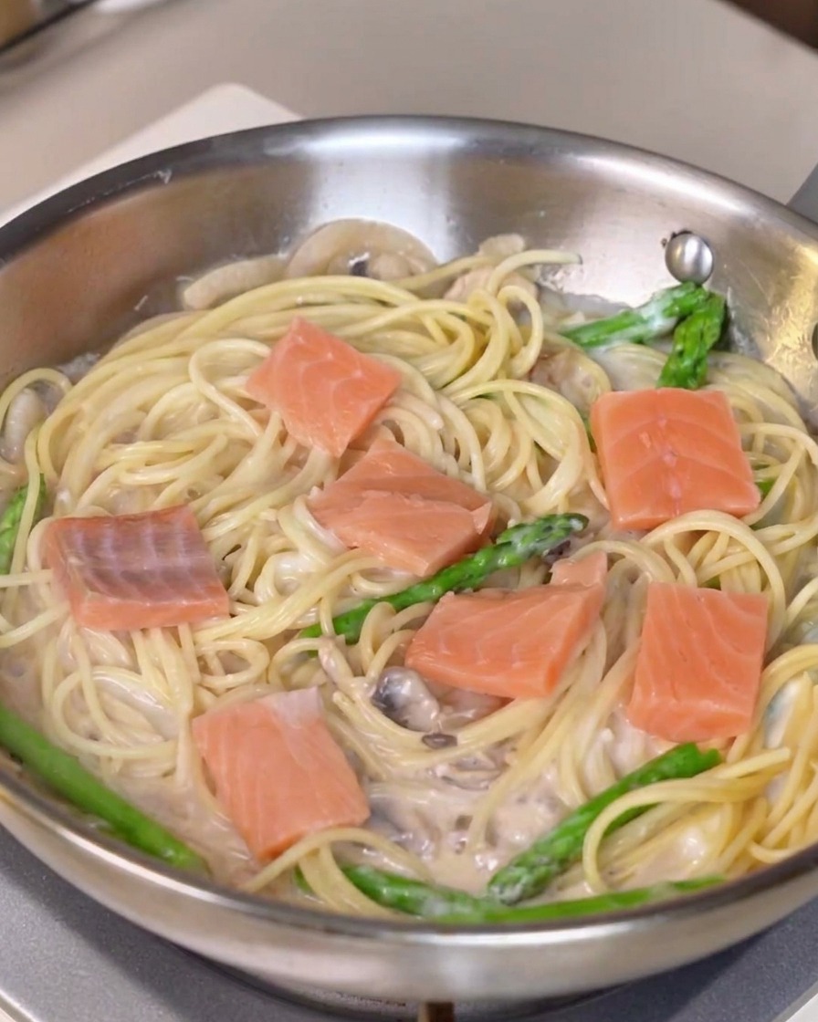 Fresh pieces of salmon being distributed over a pan of creamy pasta and asparagus.