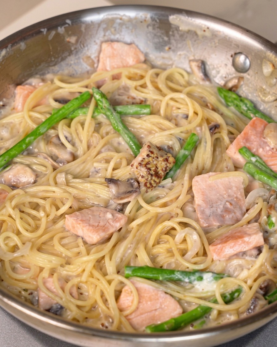 Mustard sauce, salt, and pepper being mixed into a pan of creamy salmon pasta.