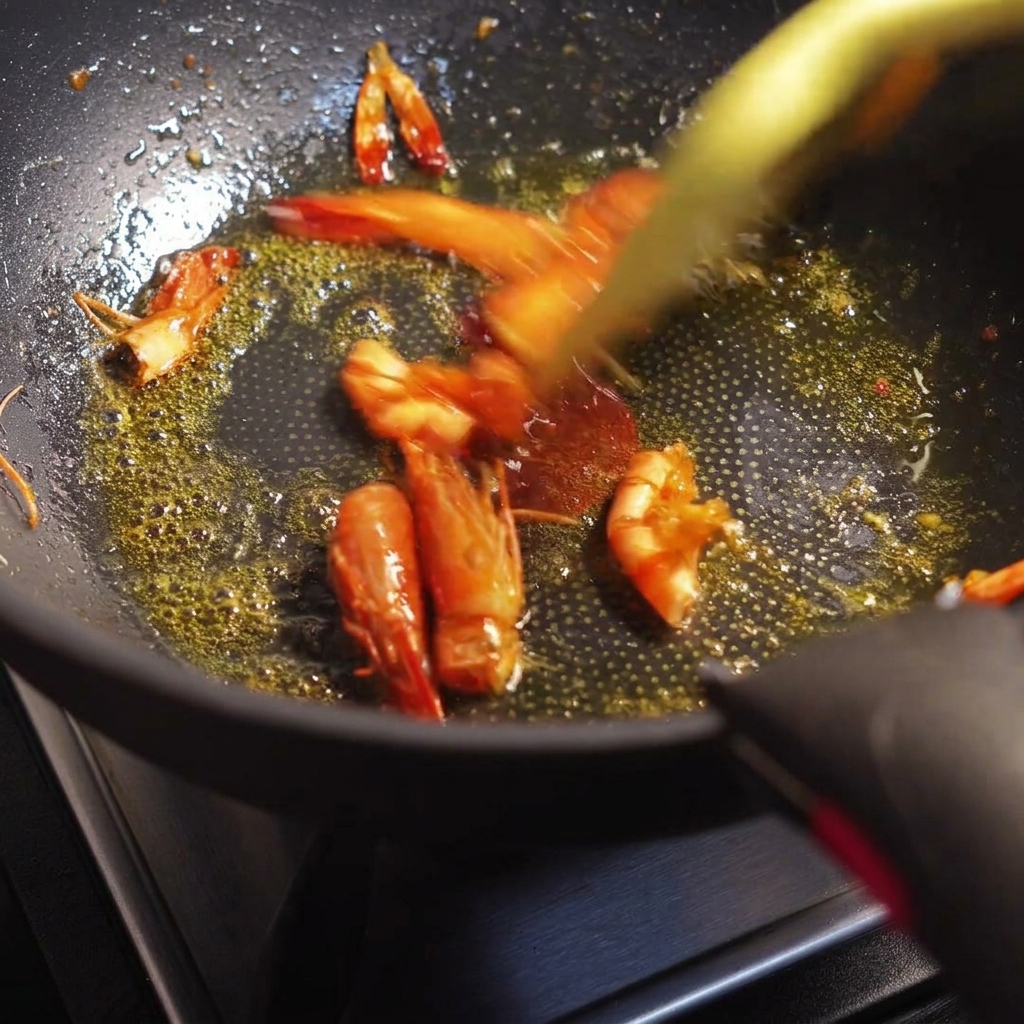 Prawn heads and shells being stir-fried in a pan, releasing a vibrant orange-red oil.