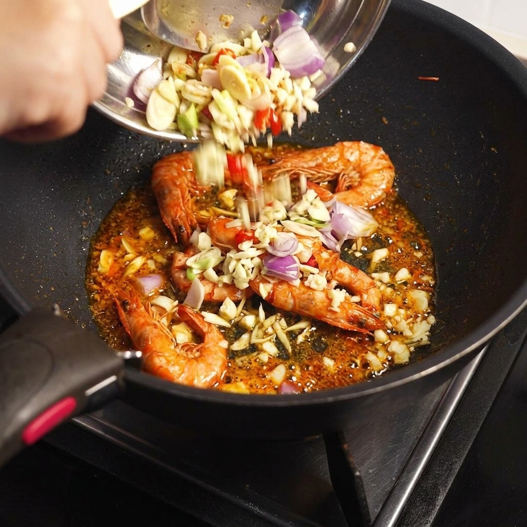 Chopped aromatics and Tom Yum paste being added from a bowl into a pan containing fried prawn shells.