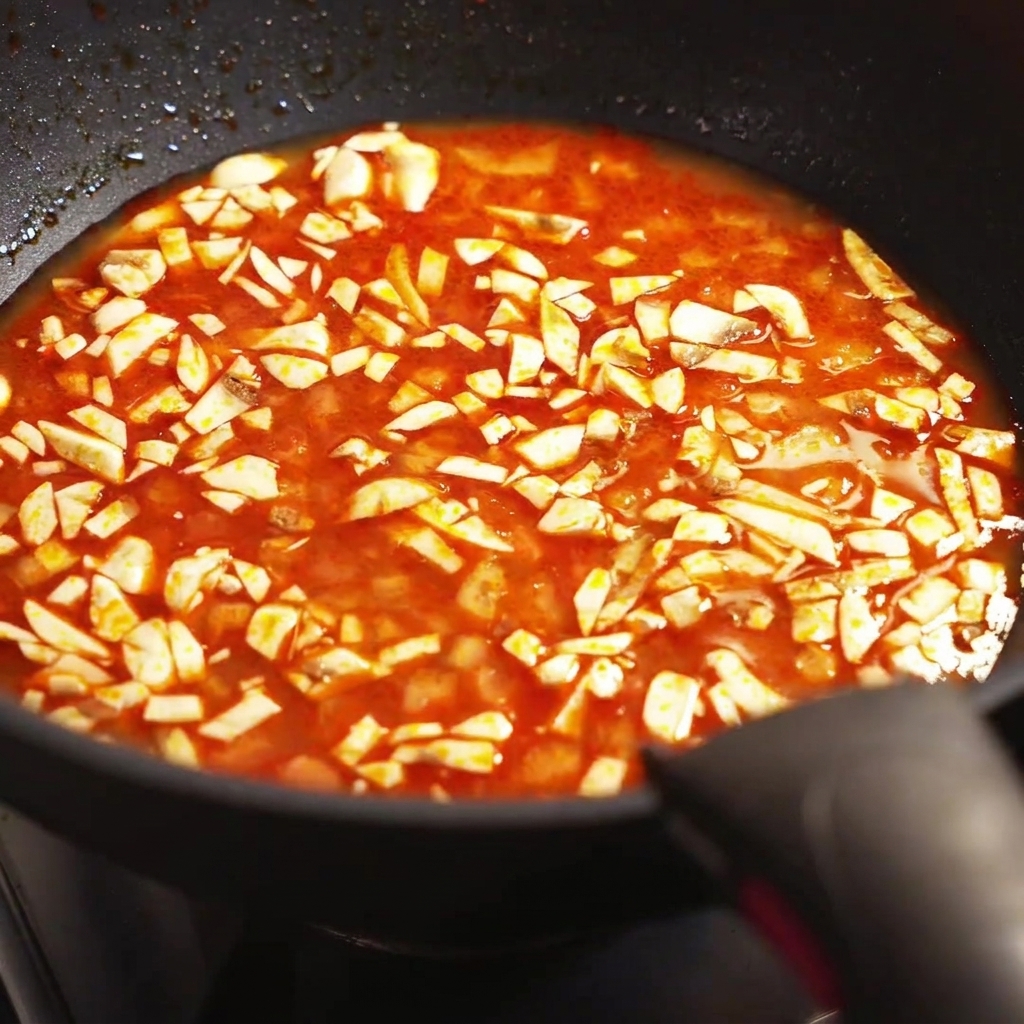 A close-up view of chopped mushrooms simmering in a seasoned red broth inside a frying pan.