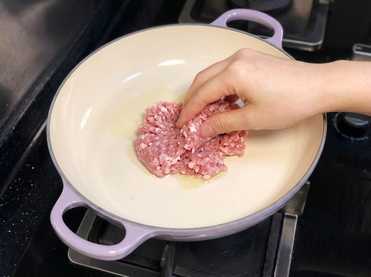 A hand placing minced pork into a purple-handled pan containing olive oil on a stovetop.