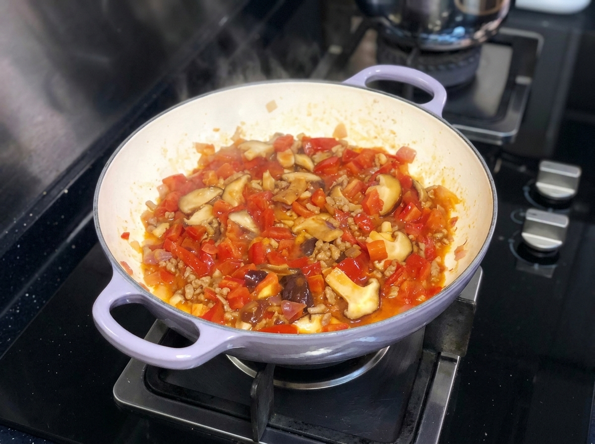 A vibrant mix of chopped tomatoes, onions, and shiitake mushrooms simmering together in a pan on a stove.