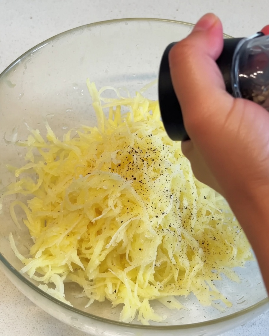 Shredded yellow potatoes inside a glass bowl receiving a seasoning of black pepper from a manual grinder.
