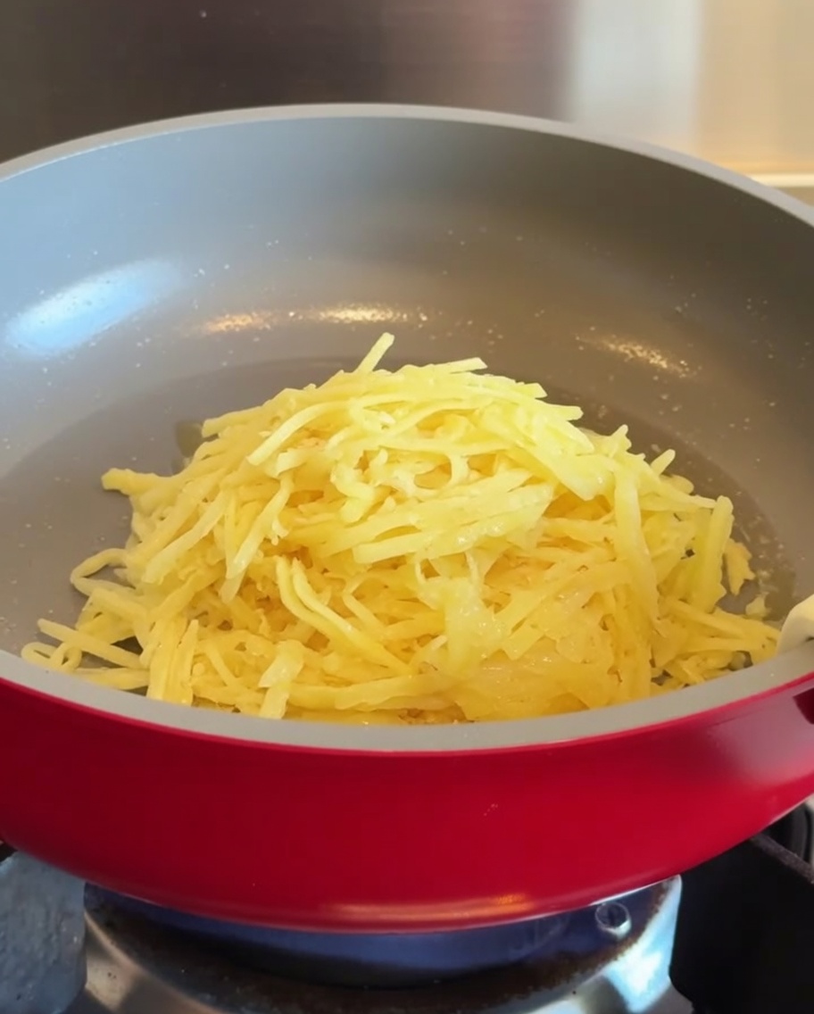 A pile of shredded potatoes placed into the center of a heated red non-stick pan.